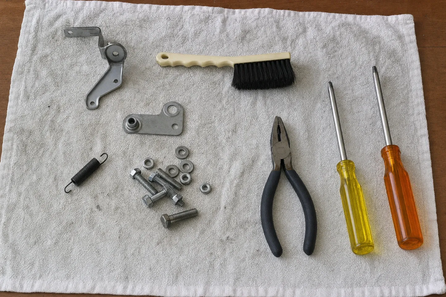Oven door hinge, a second hinge bracket, a tension spring, bolts and washers laid out on a towel beside pliers and two screwdrivers